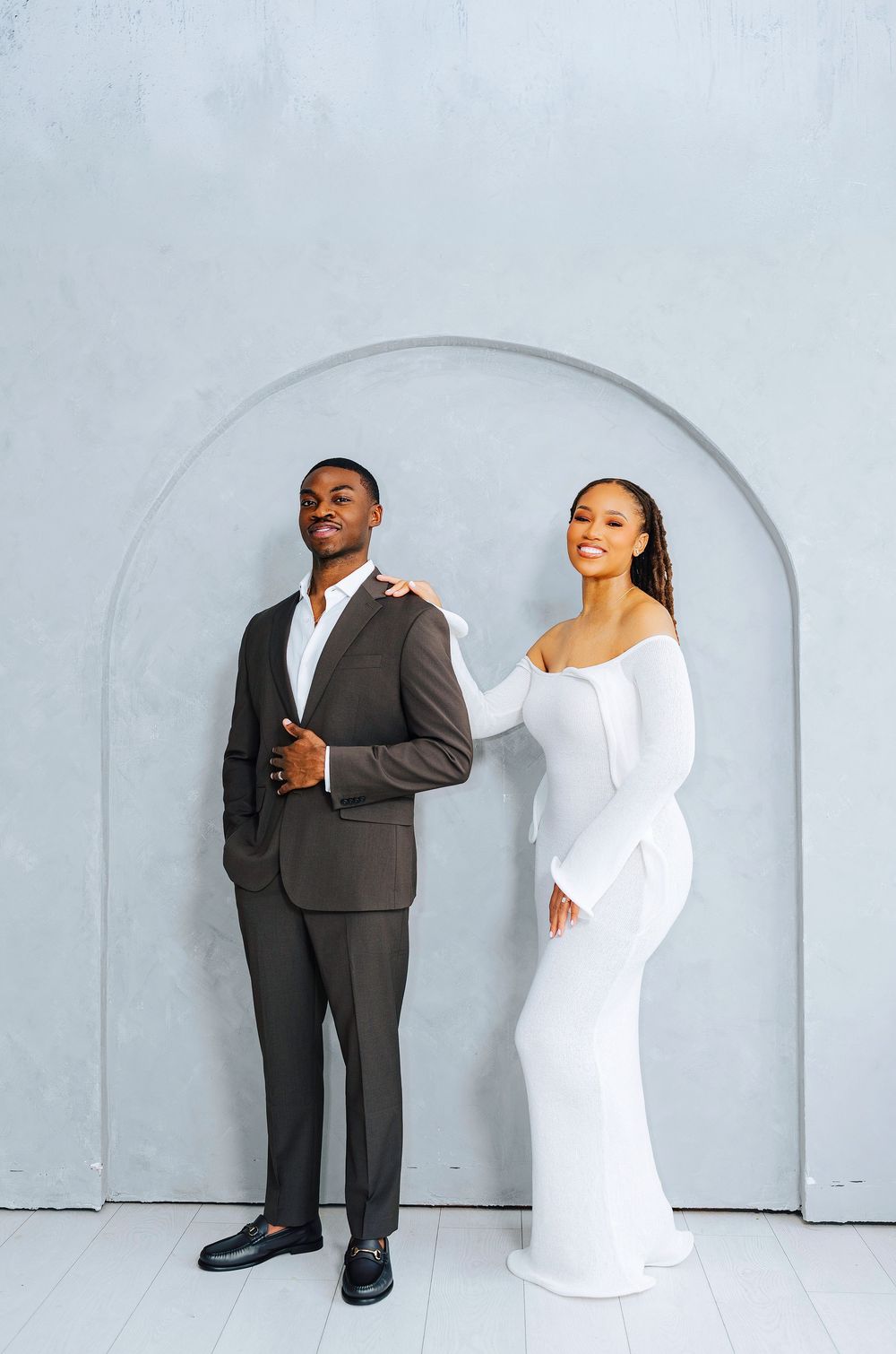 Elegant couple posing under an arch, man in dark suit and woman in white off-shoulder gown.
