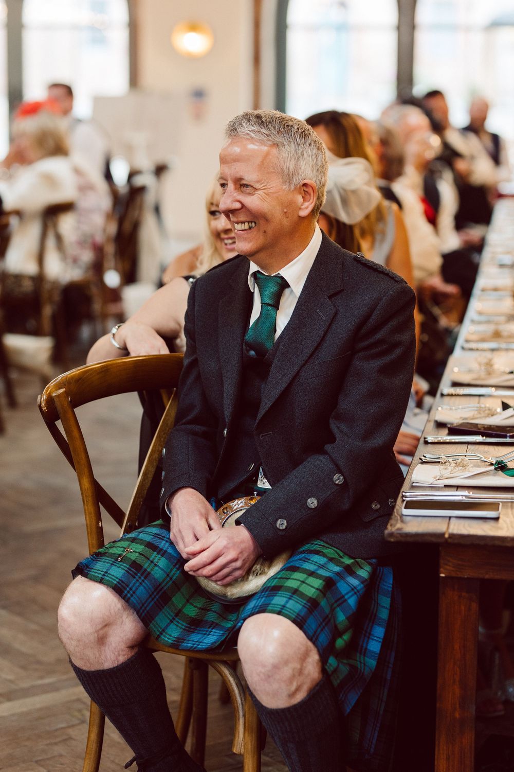 Three sequential photos show a person in a black suit jacket and blue tartan kilt seated at a formal event, laughing.
