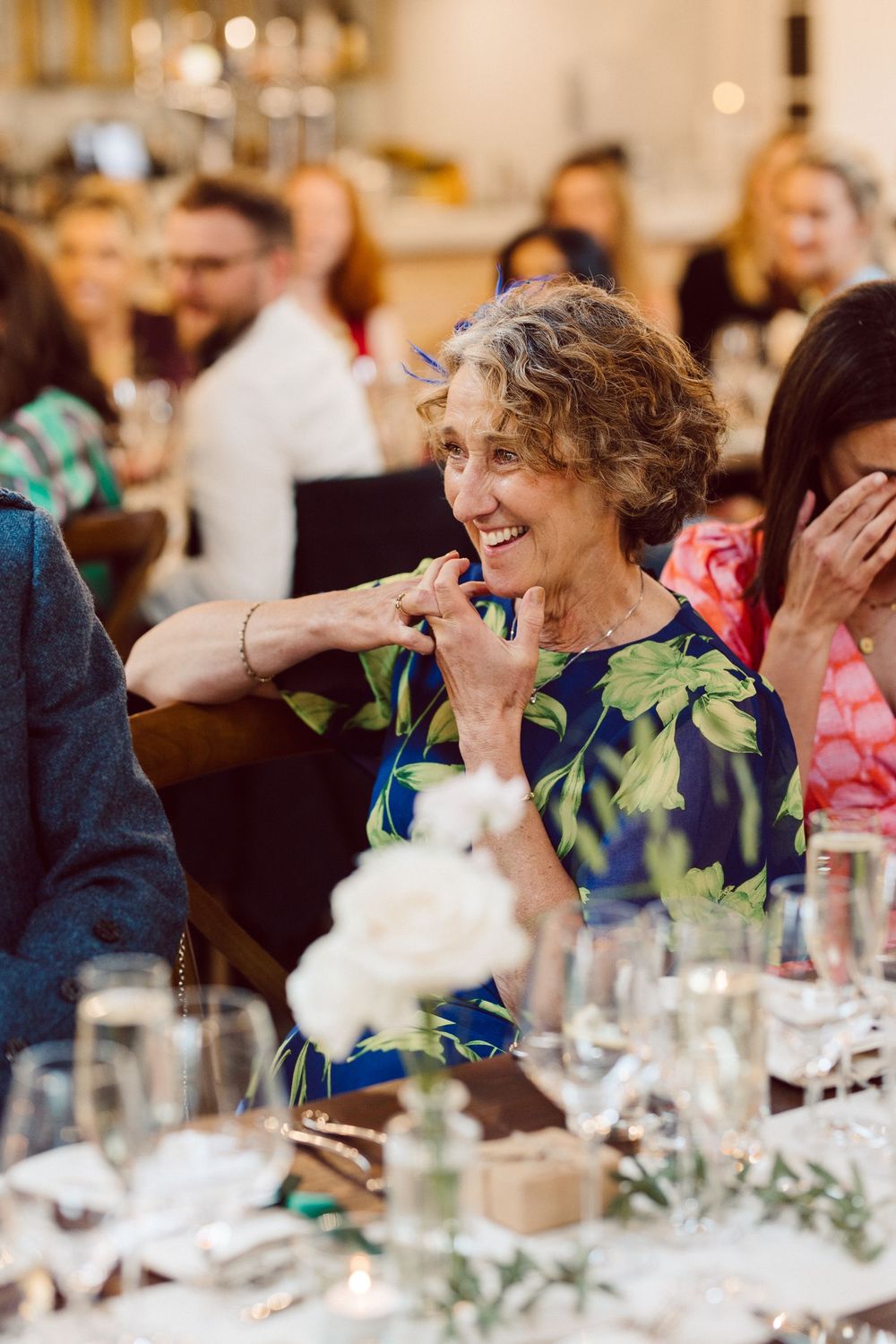 Wedding guests share joyful laughter during an elegant reception dinner with floral centerpieces and candlelight.