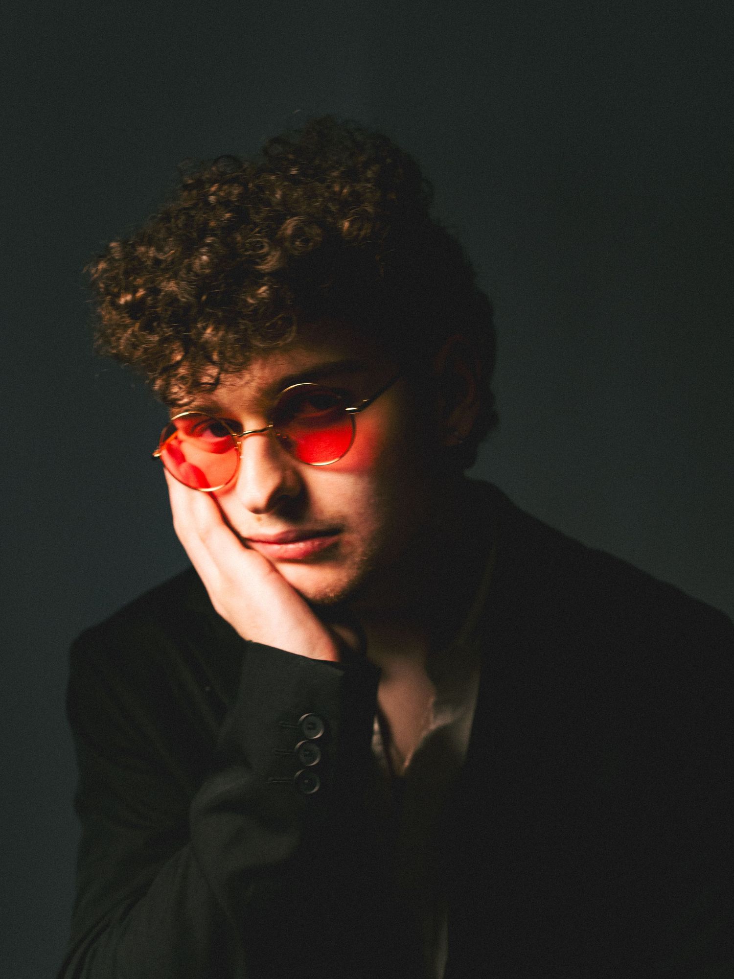 A figure in red tinted sunglasses poses against a dark background in a moody artistic portrait series.