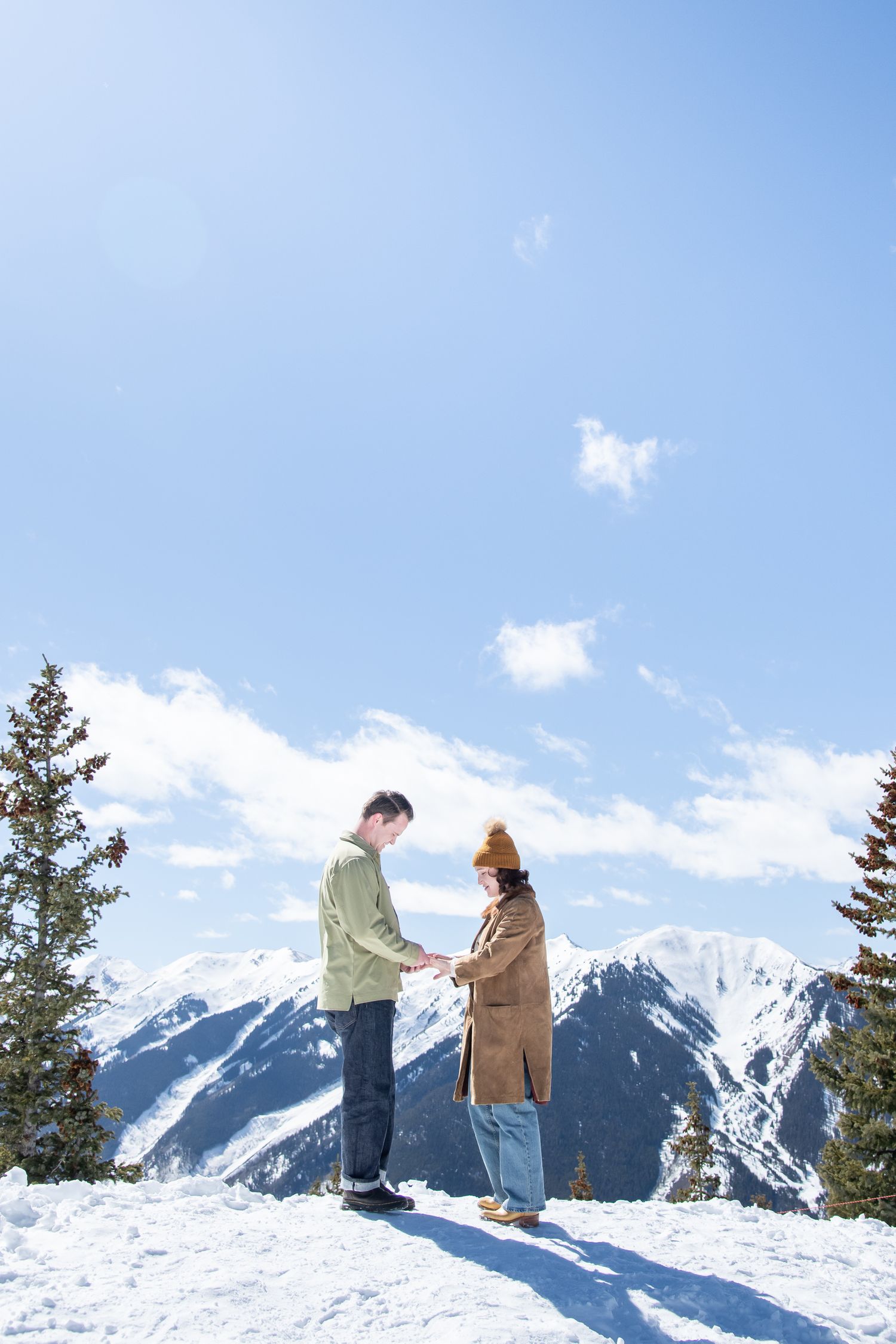 Two people share a romantic moment atop a snowy mountain peak with evergreen trees and blue skies in the background with Aspen photographer.