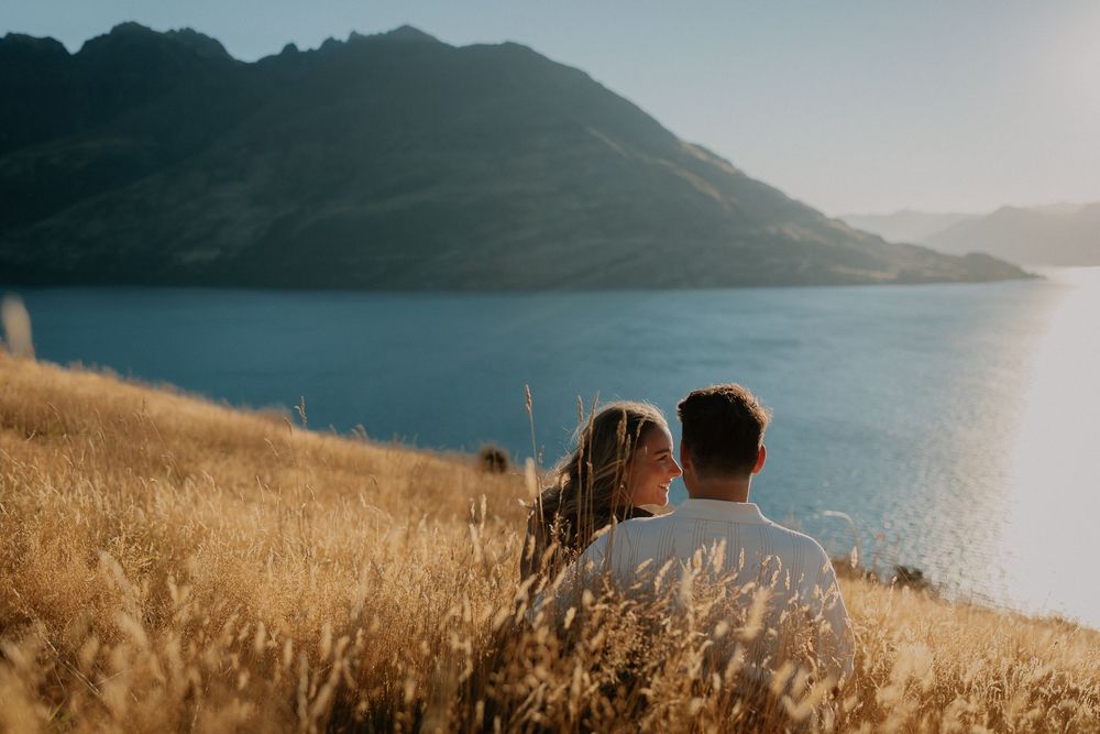 Two people sit together on a golden hillside overlooking a serene mountain lake on a sunny afternoon.