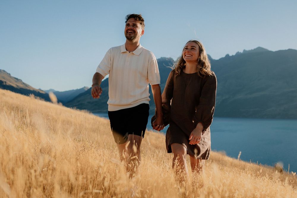 Couple walking through golden grass field at sunset with mountain views in the background.