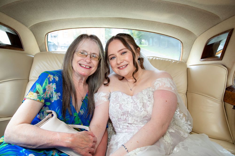 Bride in lace off-shoulder gown sits with older woman in vintage wedding car backseat smiling.