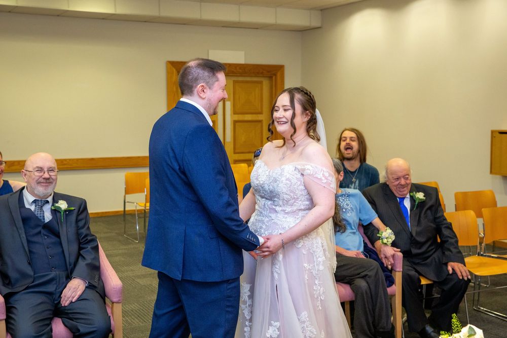 Bride and groom hold hands and smile during intimate civil wedding ceremony with seated guests.
