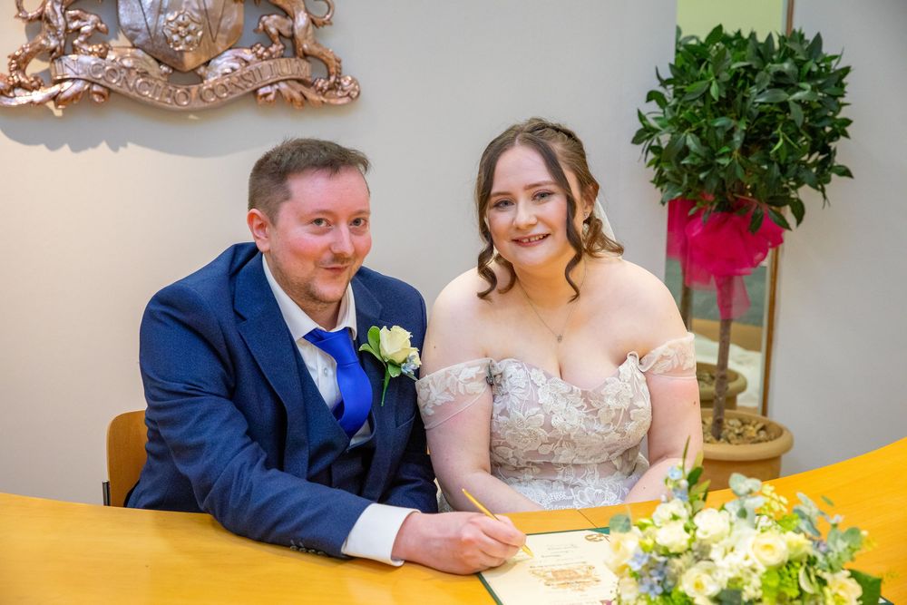 Smiling newlyweds sign wedding register at registry office table with floral bouquet nearby.