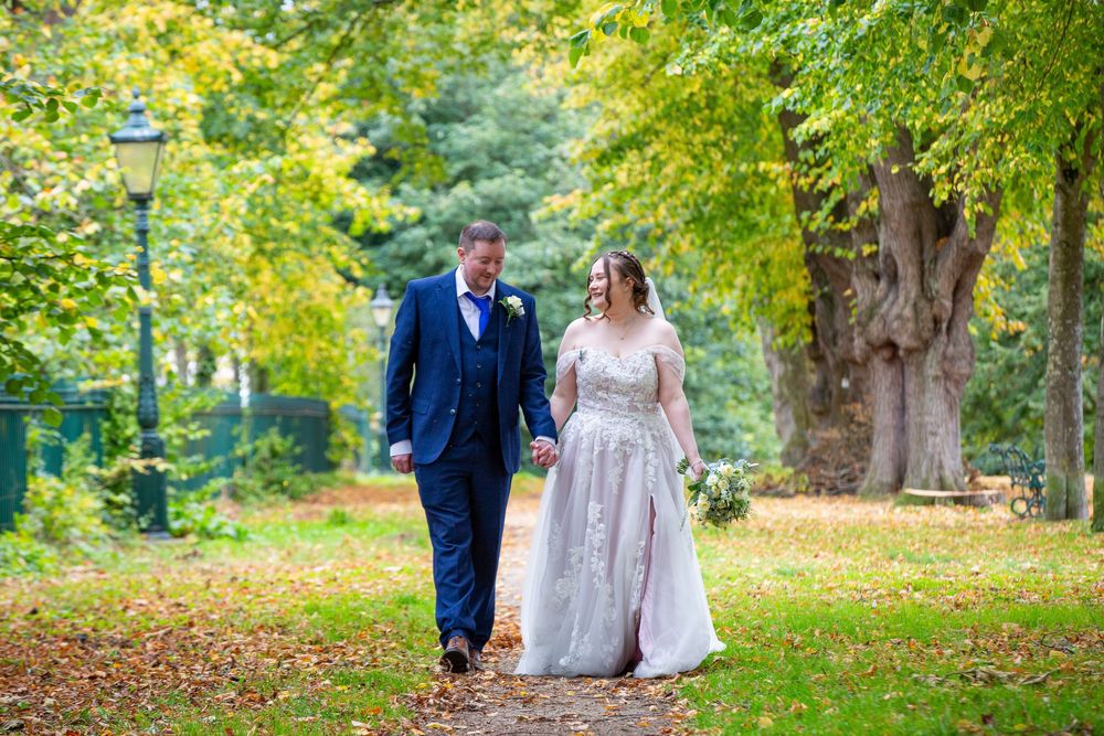 Bride and groom walk hand in hand along a tree-lined autumn path in a lush green park.