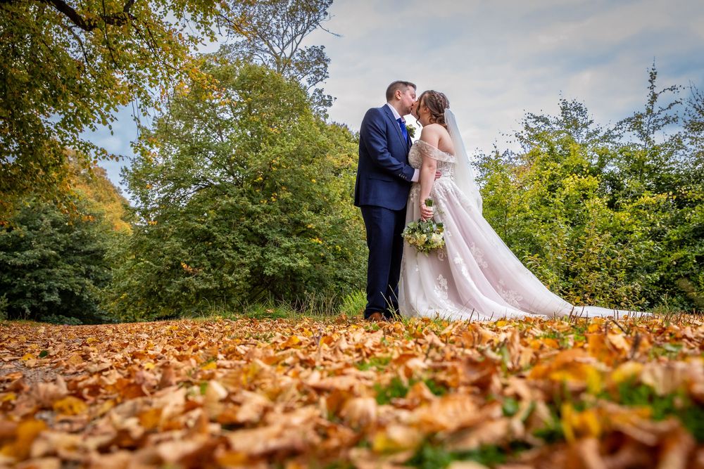 Newlyweds share a kiss surrounded by golden autumn leaves with lush green trees in the background.