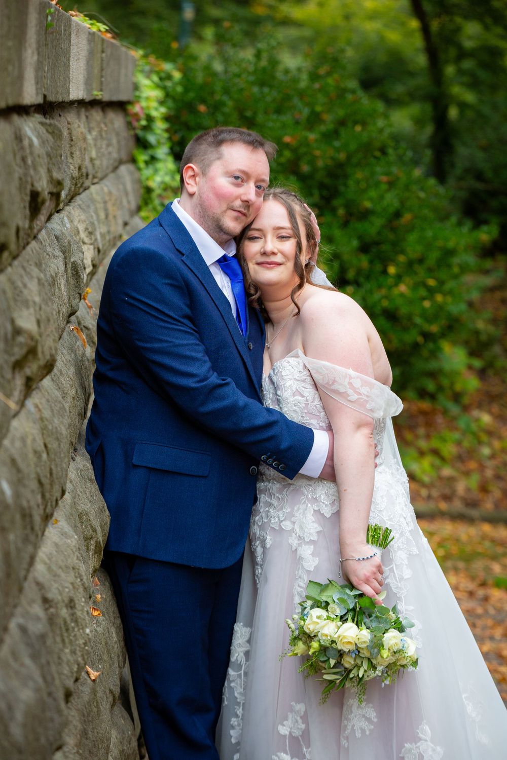 Bride and groom pose romantically against a stone wall surrounded by autumn foliage in wedding attire.