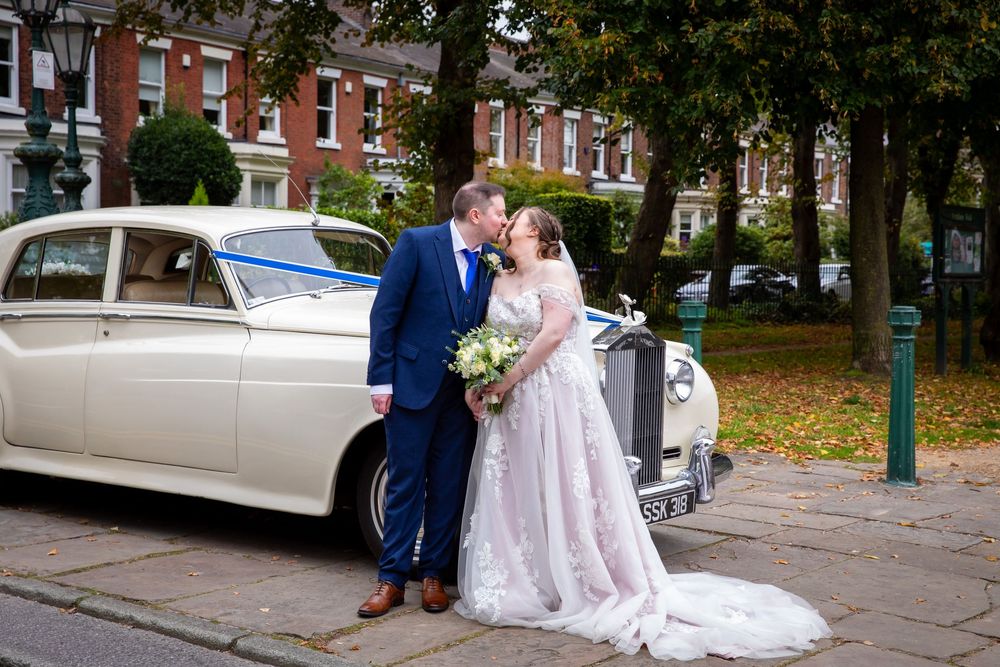 Bride and groom kiss beside a vintage white Rolls-Royce on a tree-lined street in autumn.