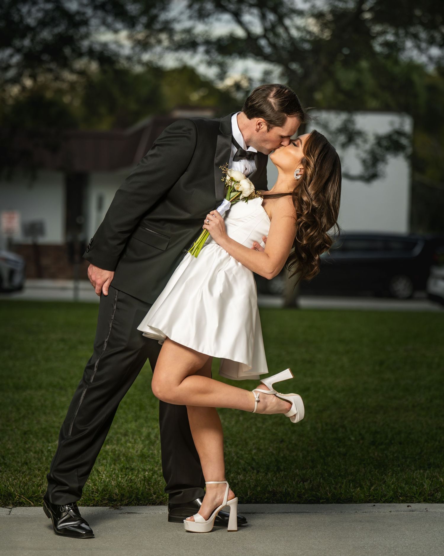 Couple sharing a romantic kiss outdoors, groom in black tuxedo and bride in short white dress holding a bouquet.