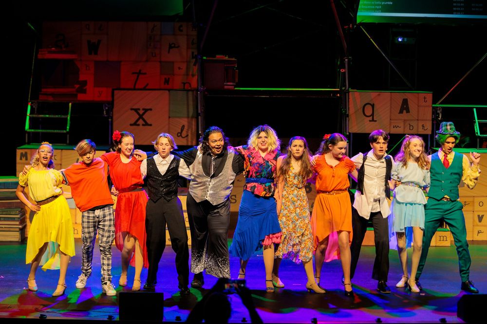 A full cast of young performers takes a bow on a colorful stage set at the end of a musical production.