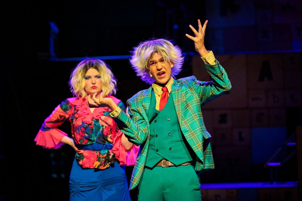 Two colorfully costumed performers on stage, one in plaid suit and pink top, during a vibrant theatrical production.