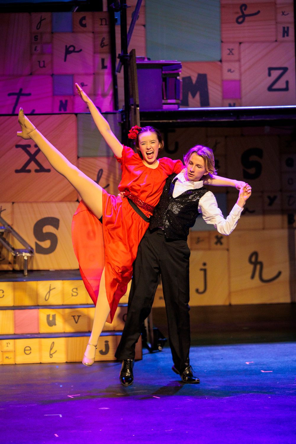 Female dancer in red dress performs high kick lift with male partner on a letter-decorated theatrical stage.