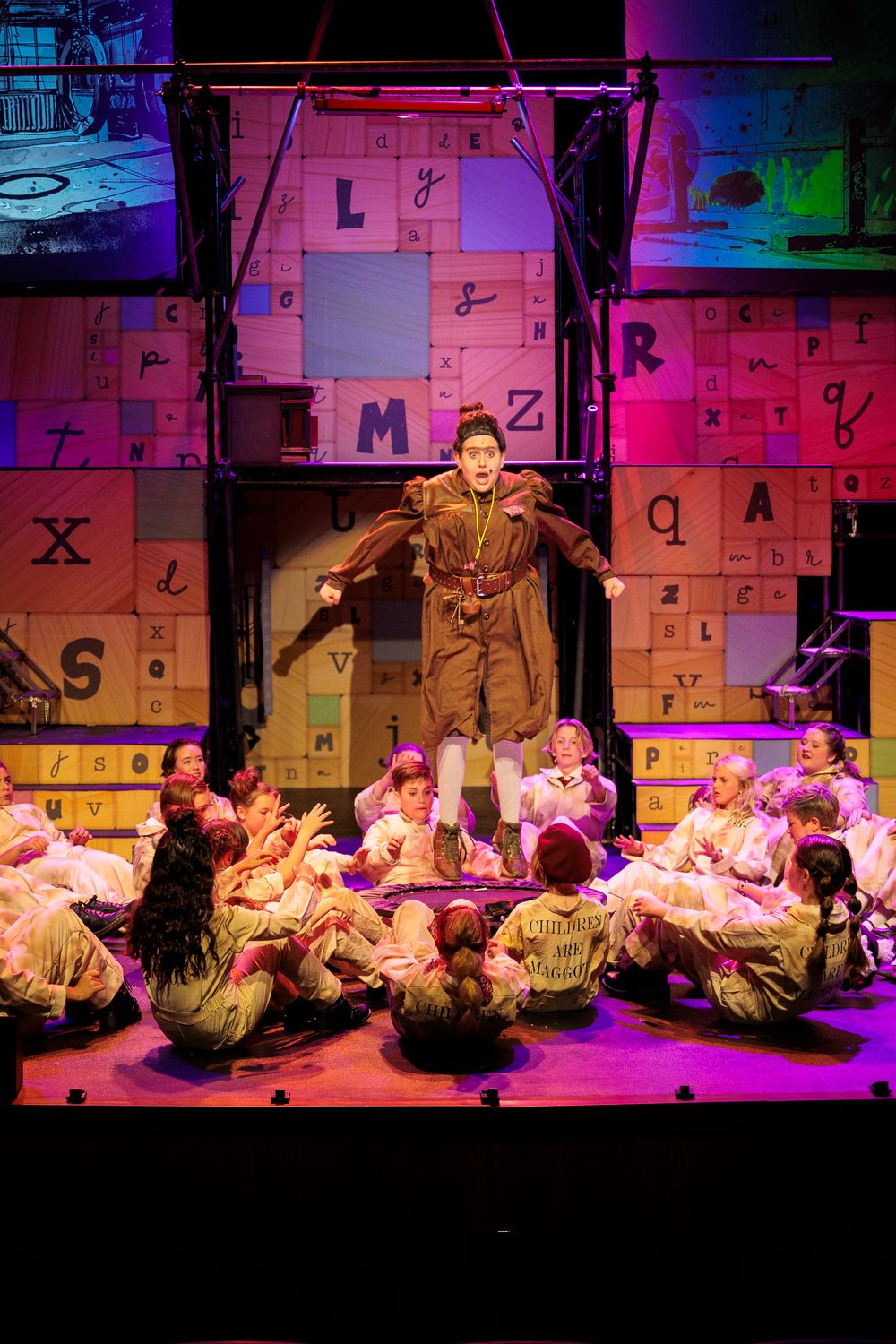 Performer leaps above seated children in matching costumes on a letter-themed stage set during a theatrical show.