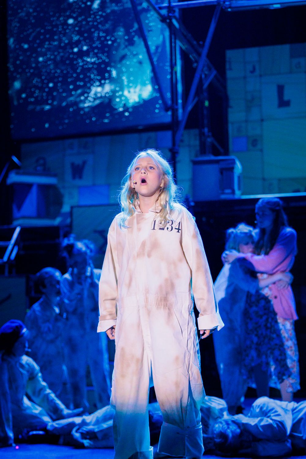Young performer in white numbered costume sings solo under blue stage lighting in an emotional theater scene.