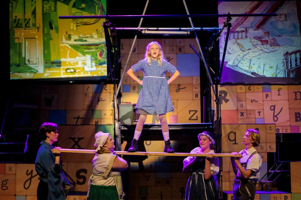 Young girl in blue dress stands elevated on stage set while surrounded by cast members in a theater production.