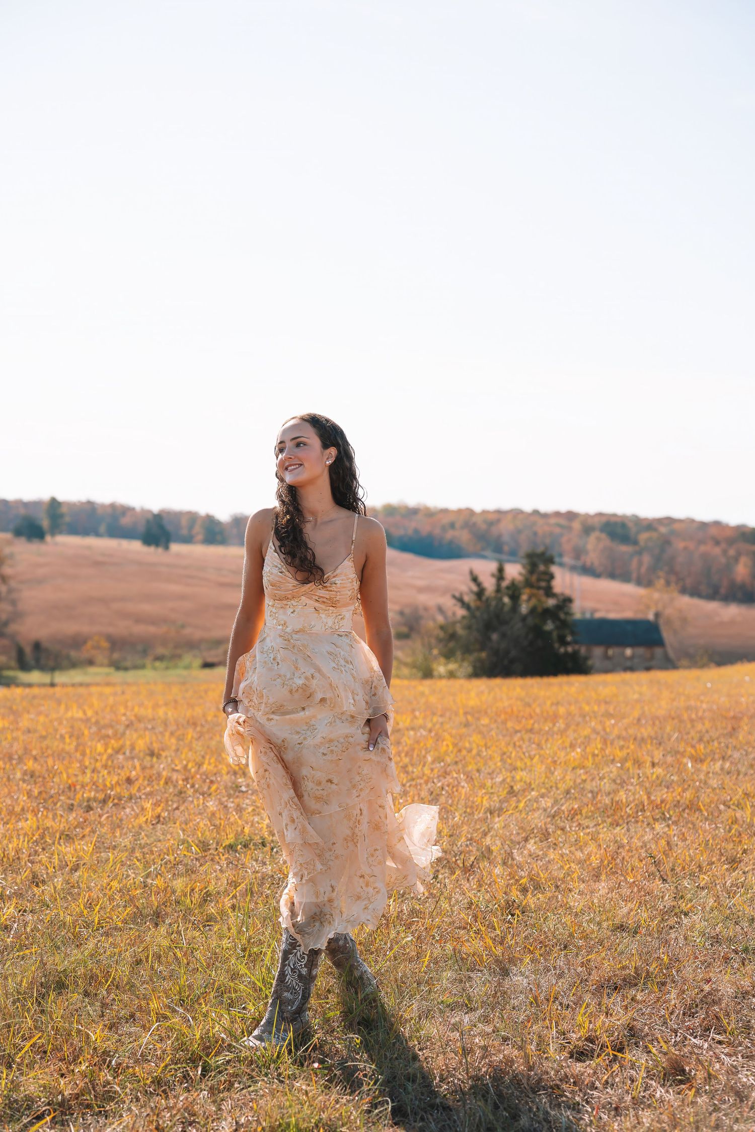 A woman wearing a floral dress stands in a field of yellow flowers, with a wooded area in the background.