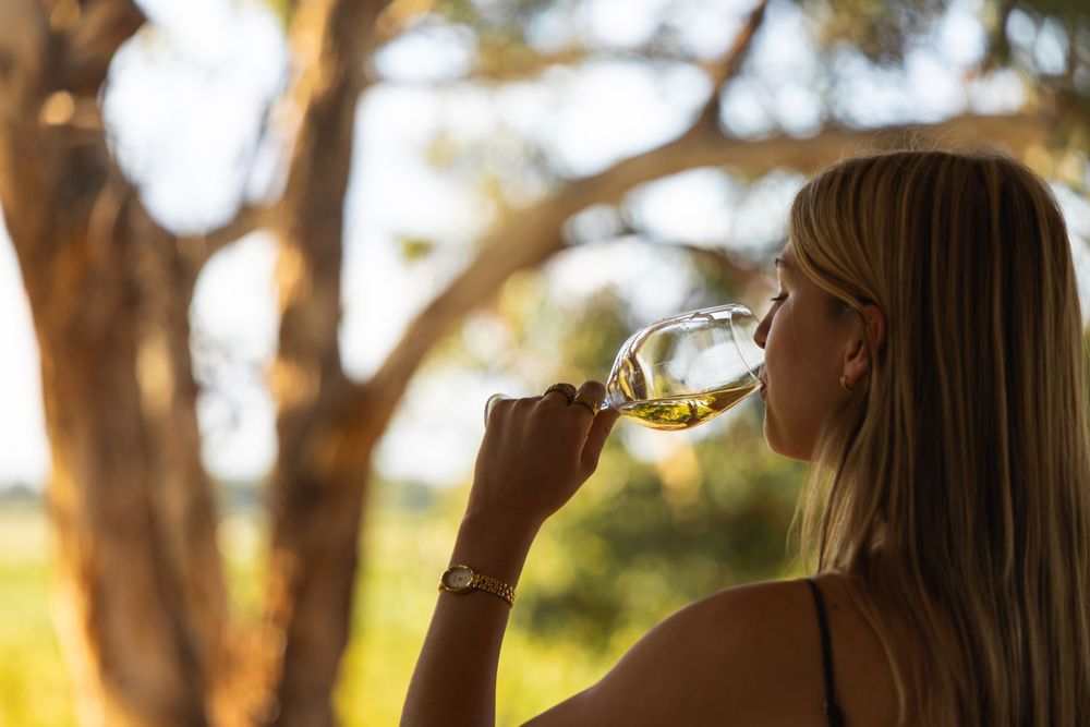 Woman drinking white wine outdoors with vineyard and trees in background