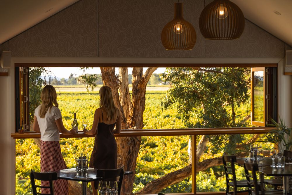 Two women standing inside winery with vineyard views through open windows