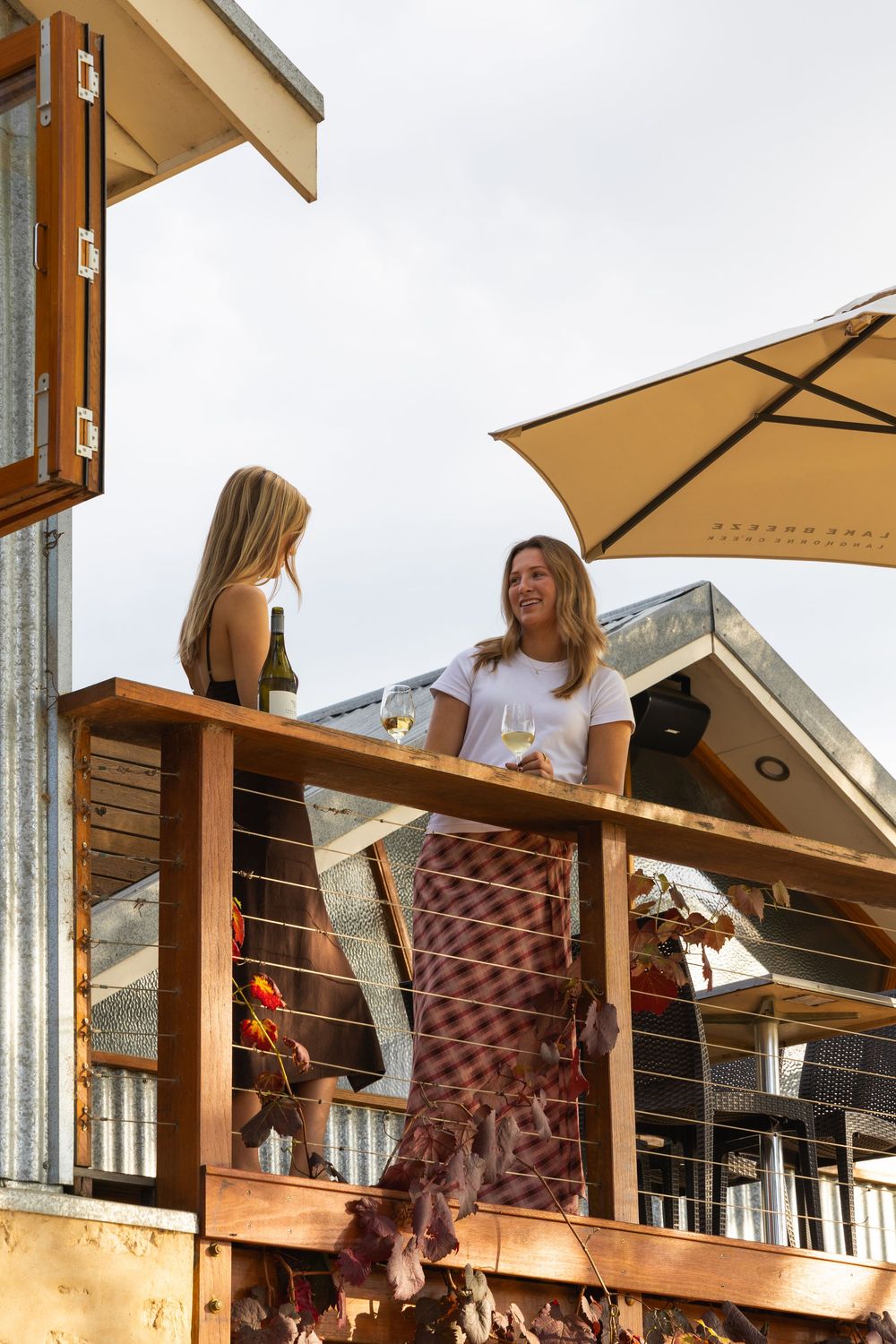 Two women talking on winery balcony under umbrella with modern building exterior