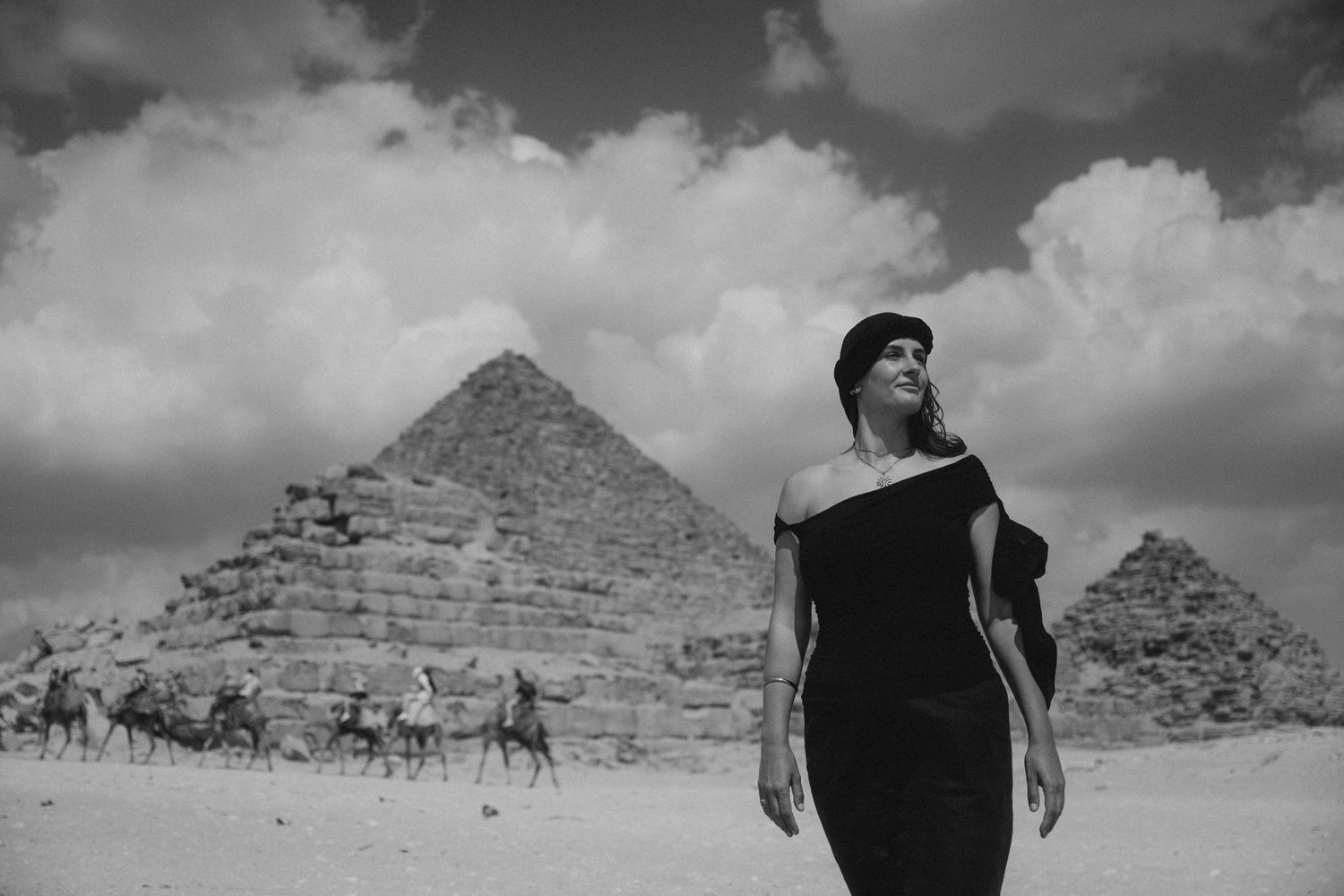 A dramatic black and white panoramic view of tourists visiting the ancient pyramids of Giza in Egypt under cloudy skies.