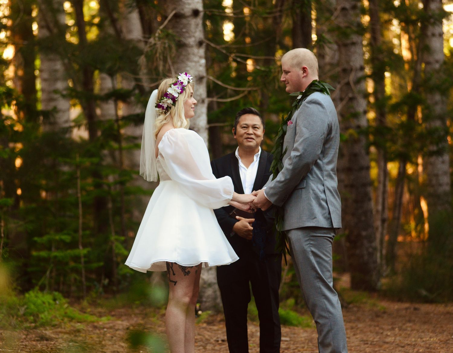 A couple exchanges wedding vows during an outdoor forest ceremony with golden sunlight filtering through the trees.