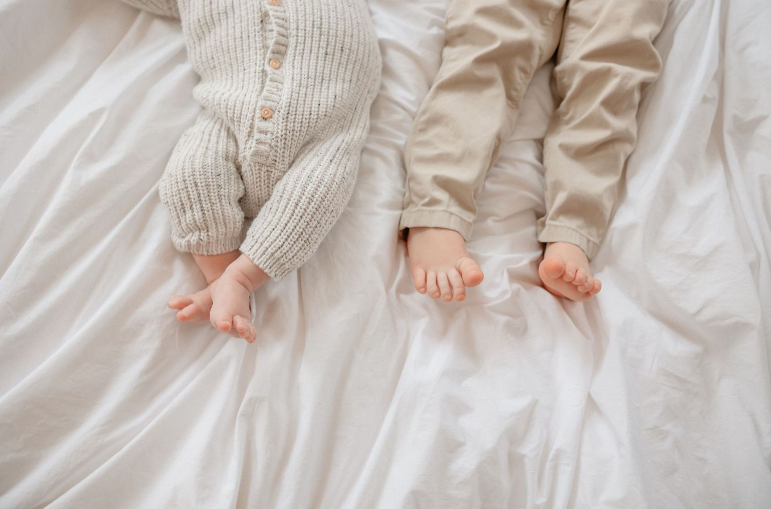 Tiny baby feet wearing white knit booties on a soft white bedding surface.