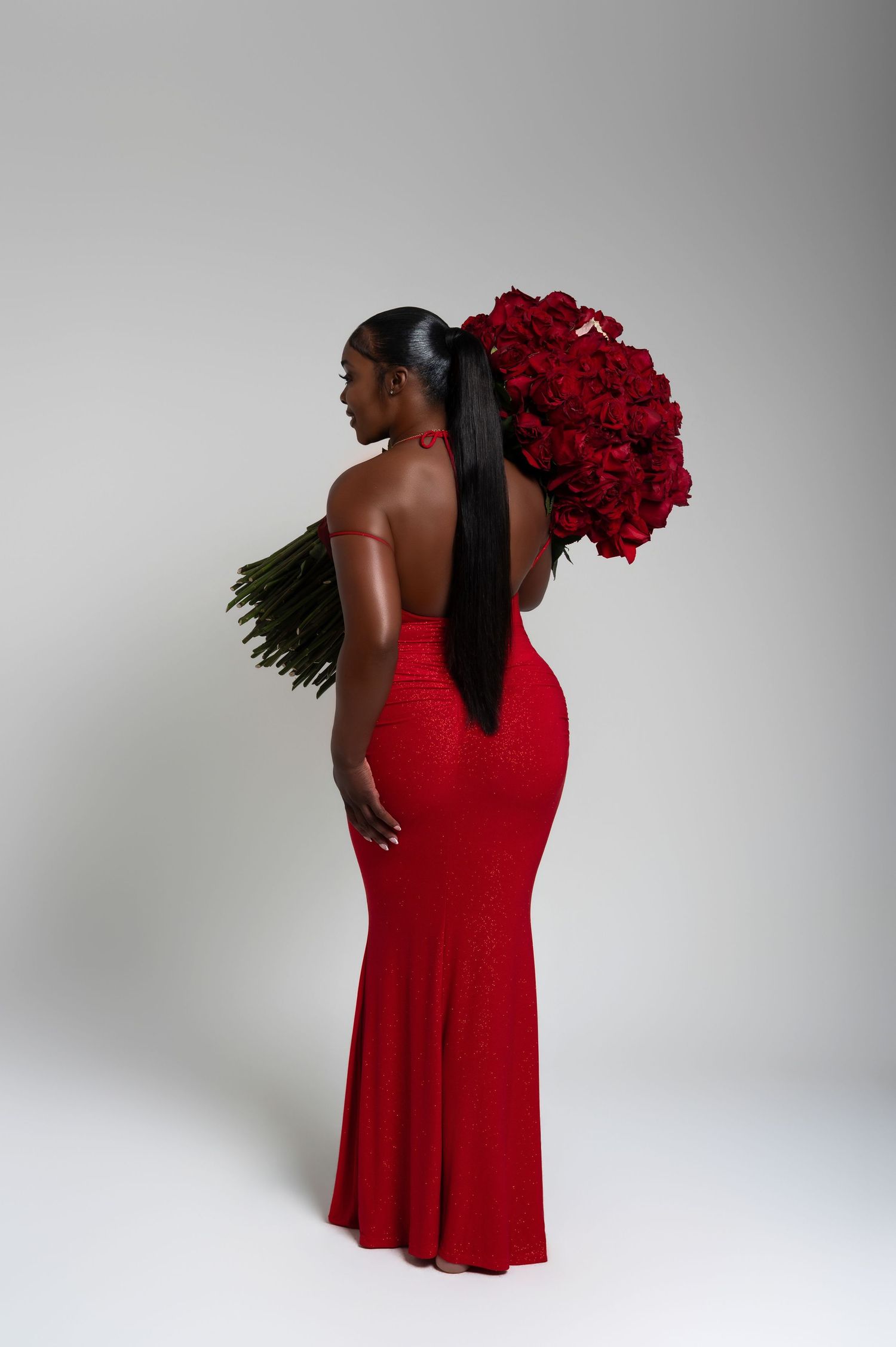 A Black woman in a red mermaid gown holds a large bouquet of red roses, posing with her back to the camera.