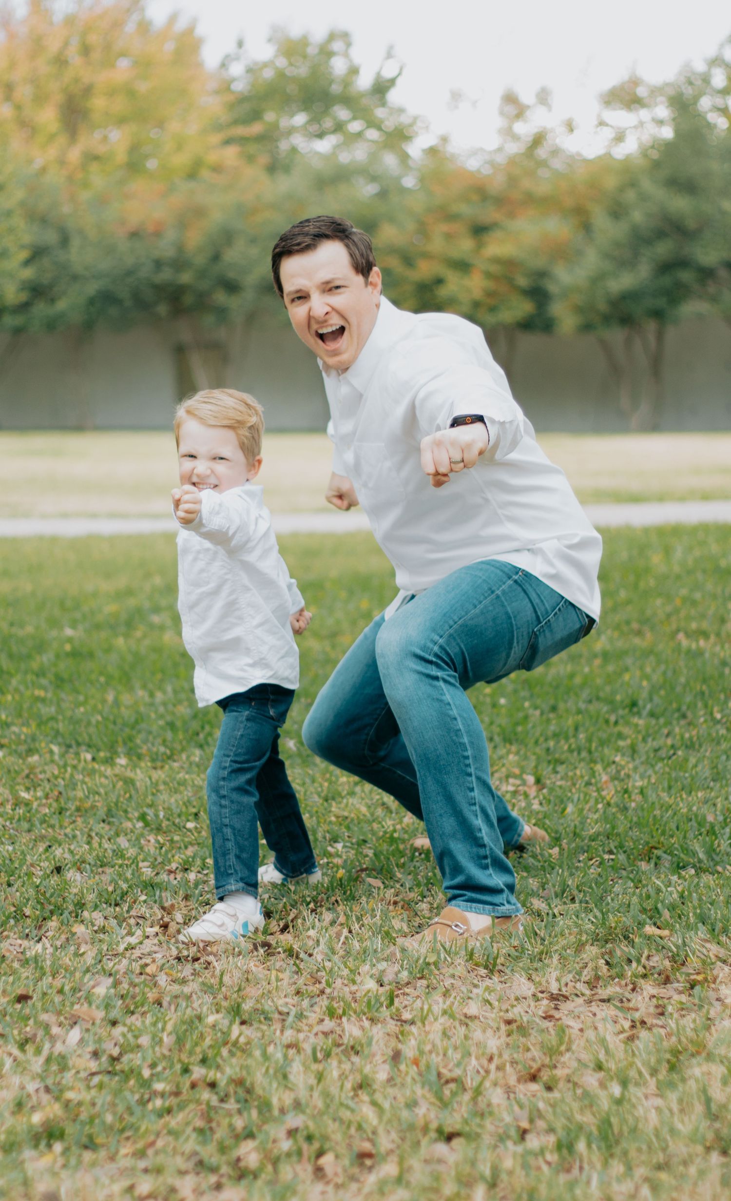 Two people in white shirts and jeans dance and play together joyfully on grass with autumn trees in the background.