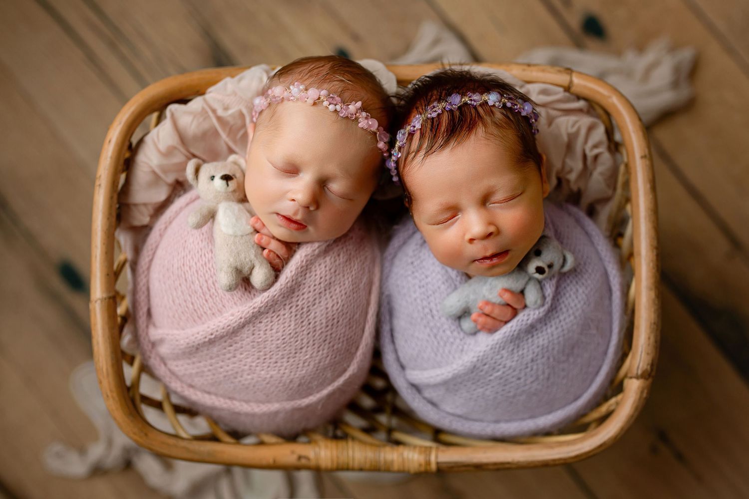 Newborn twins in pink and purple wraps and headbands with teddy bears in basket on hardwood floor in Summerville, SC
