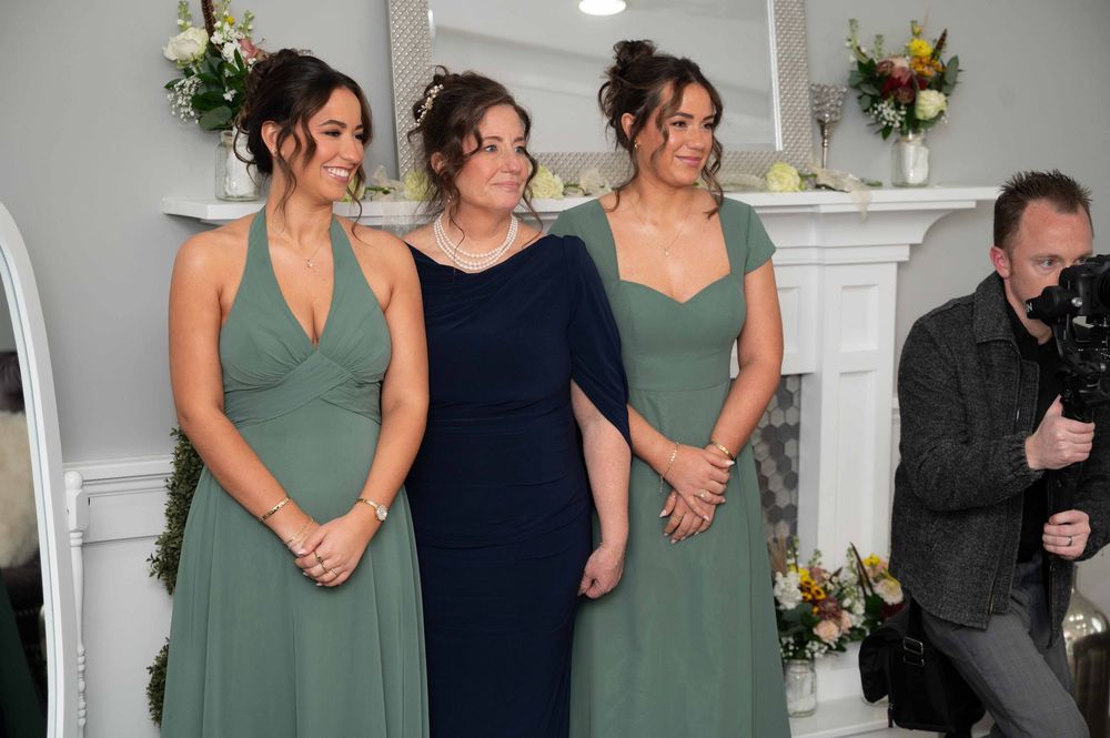 Three women in formal attire stand together at a wedding ceremony with floral decorations and a white mantle in the background.