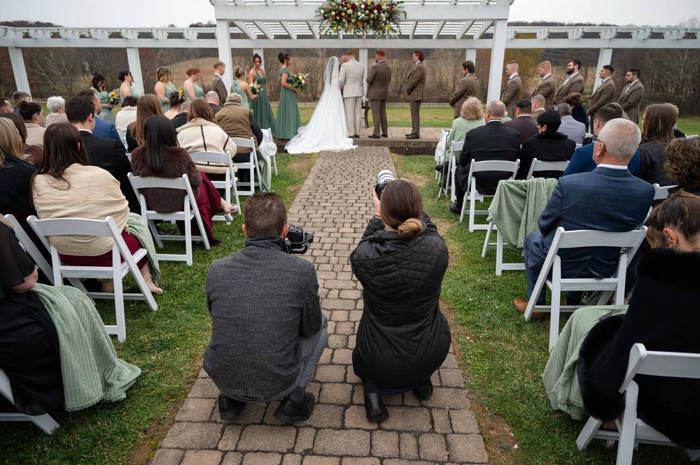 Wedding guests seated in white chairs during an outdoor ceremony on a stone patio and grassy area.