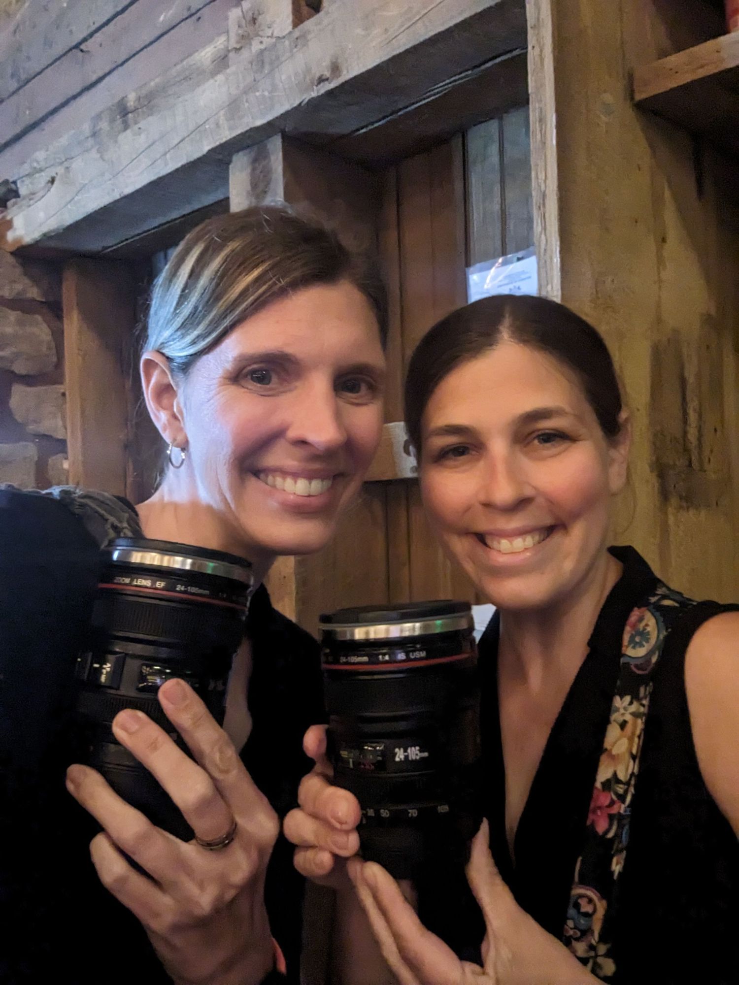 Two photographers hold up their professional Canon camera lenses while posing together indoors.