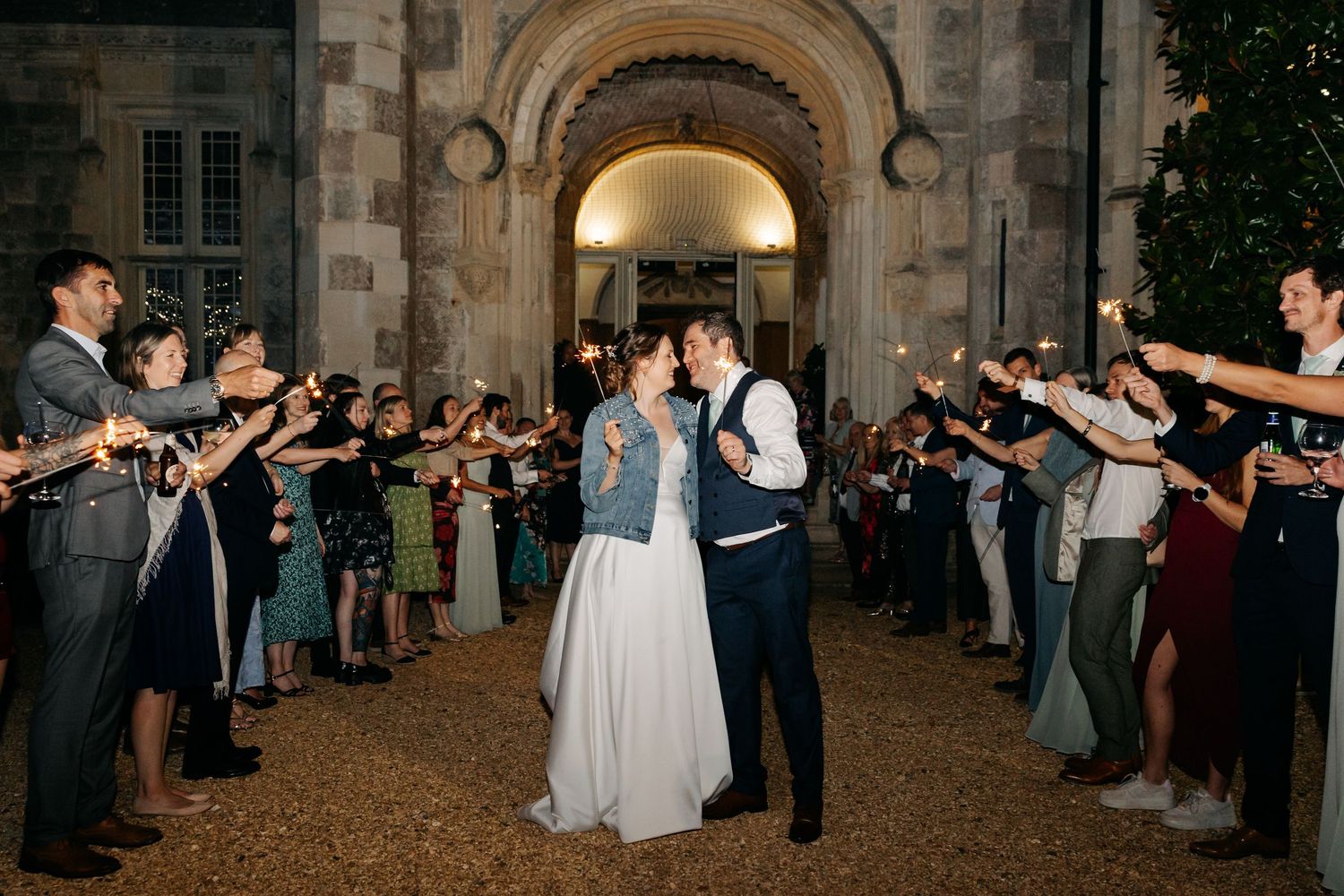 Wedding guests form a sparkler tunnel exit at night outside a stone archway as newlyweds depart their celebration.