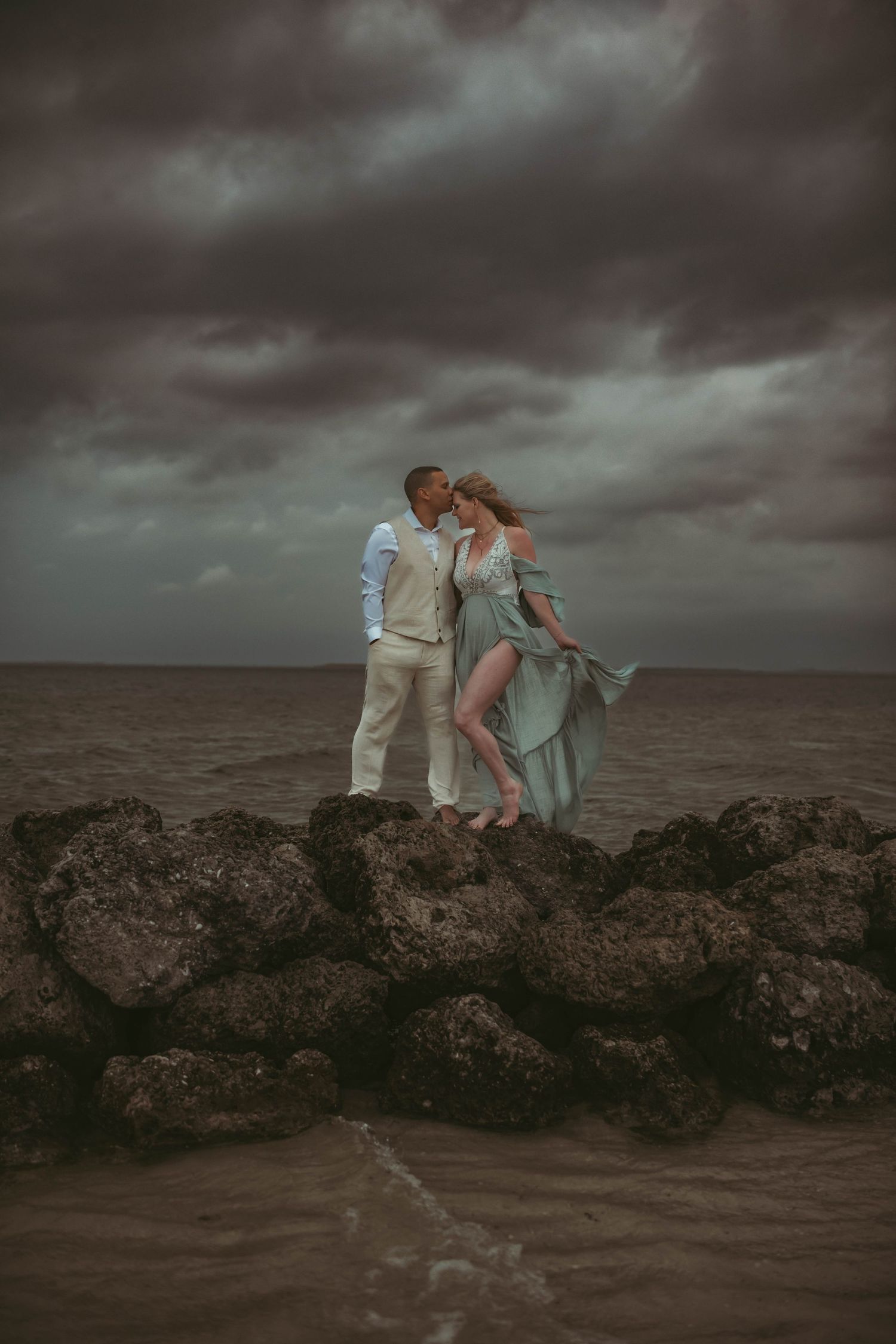 A couple stands on rocky coastal cliffs at dusk under dramatic stormy skies.