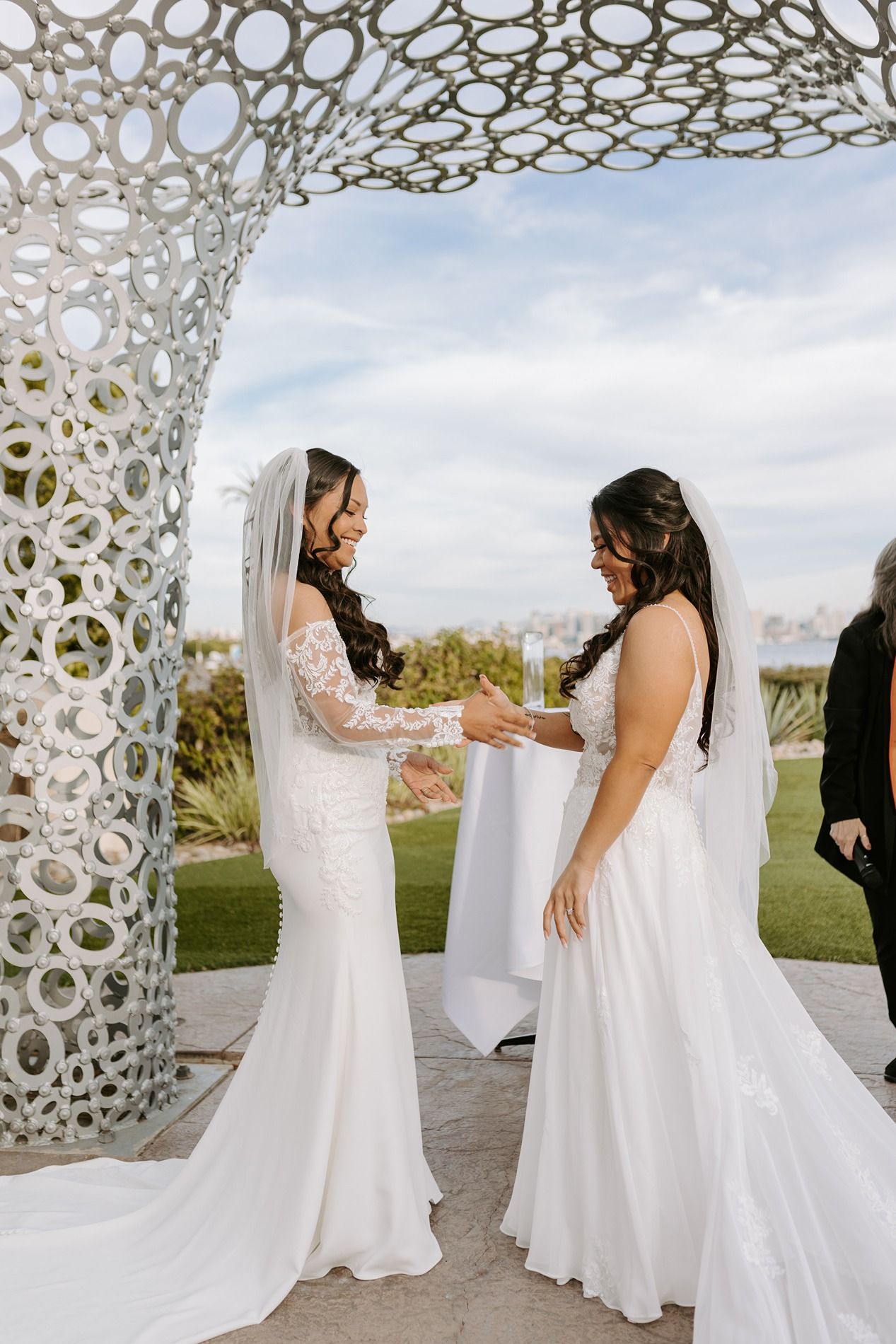 Two brides in flowing white wedding gowns share a joyful moment under a decorative white archway outdoors.