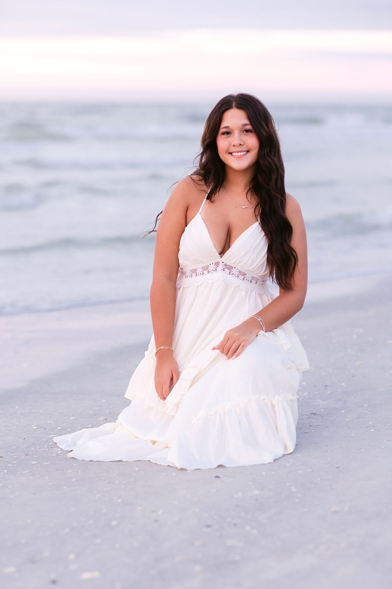 Dark haired senior girl wears a white dress and poses sitting on Indian Rocks Beach at sunset in Clearwater, Florida for senior photos.