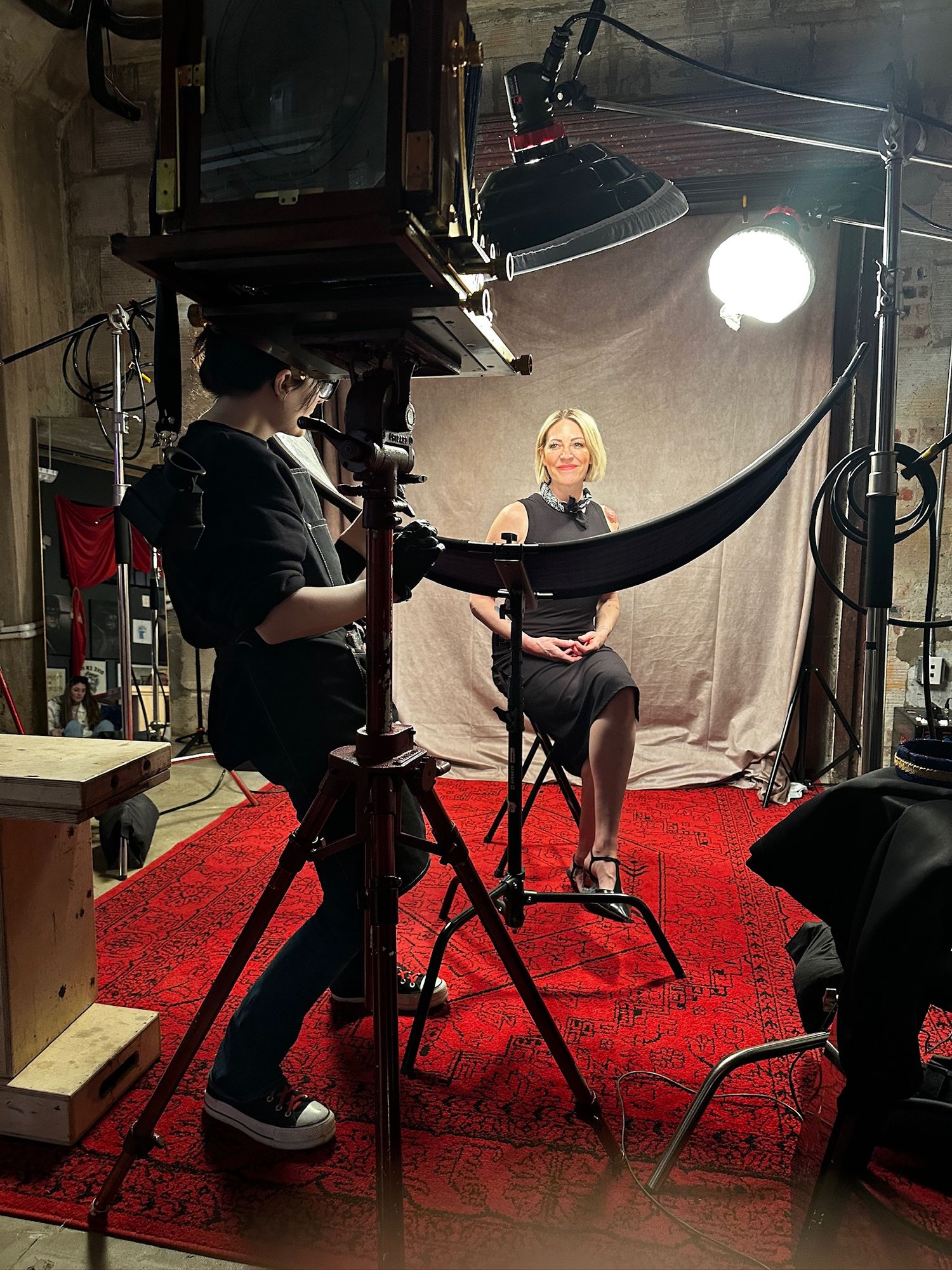 Behind the scenes photo shoot setup with lighting equipment and red carpet against white backdrop in studio.