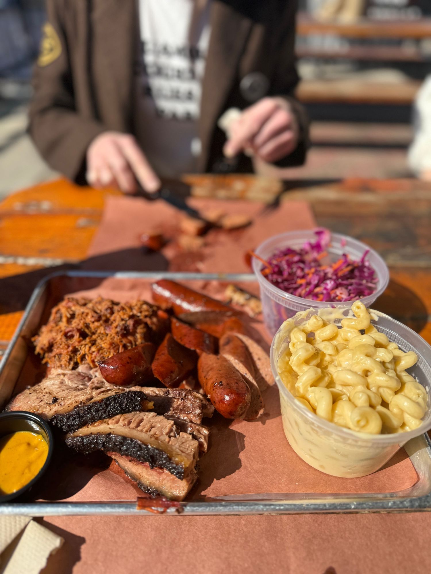 A wooden serving board displays sliced smoked brisket with sides of mac and cheese, coleslaw, and barbecue sauce.