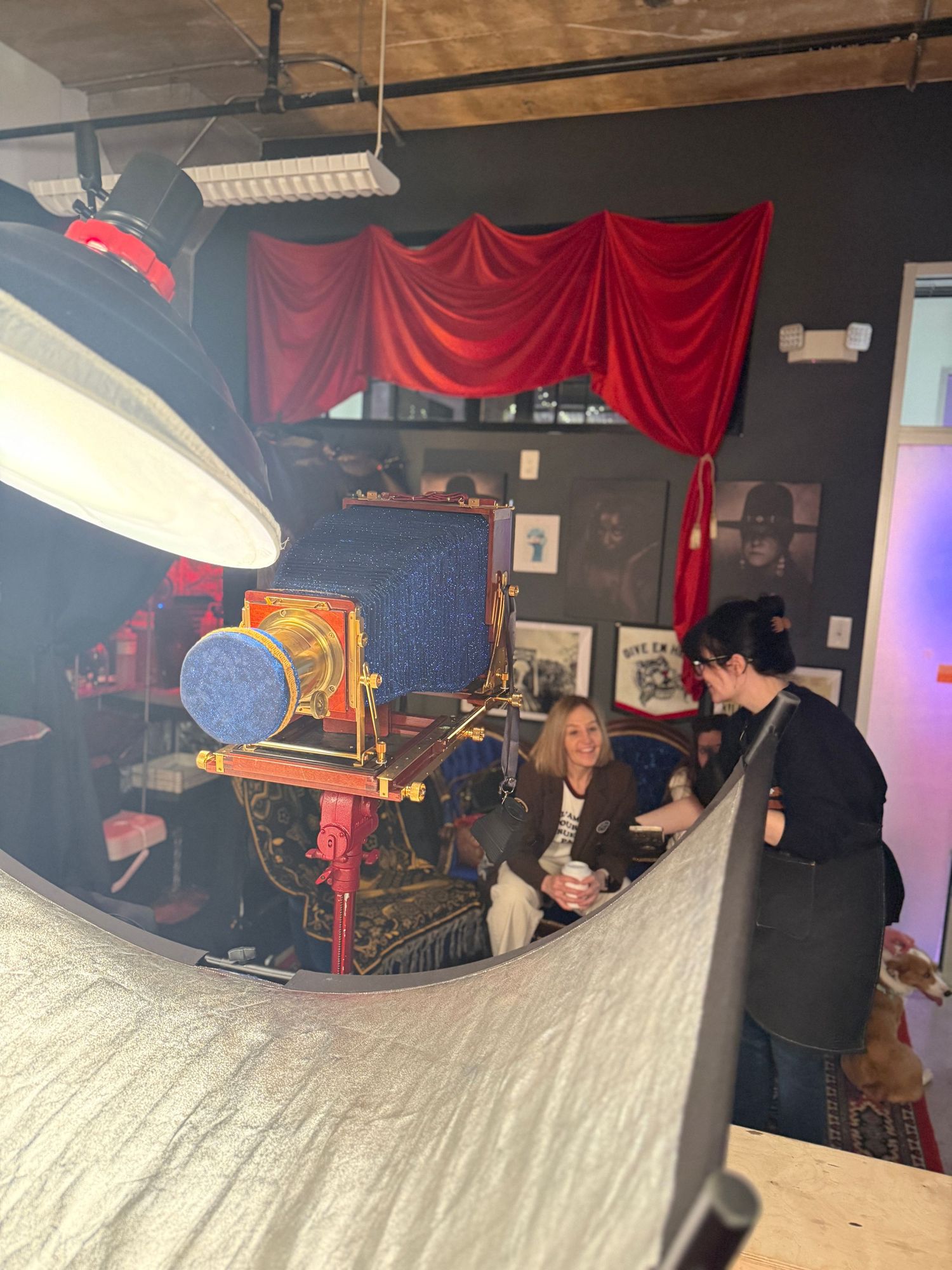 Behind-the-scenes photo of a film set with red curtains, lighting equipment, and crew members working in a dark room.