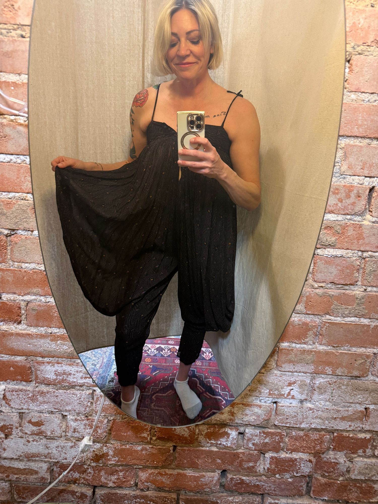 Mirror selfie showing someone wearing a black flowing jumpsuit and sneakers against a brick wall background.
