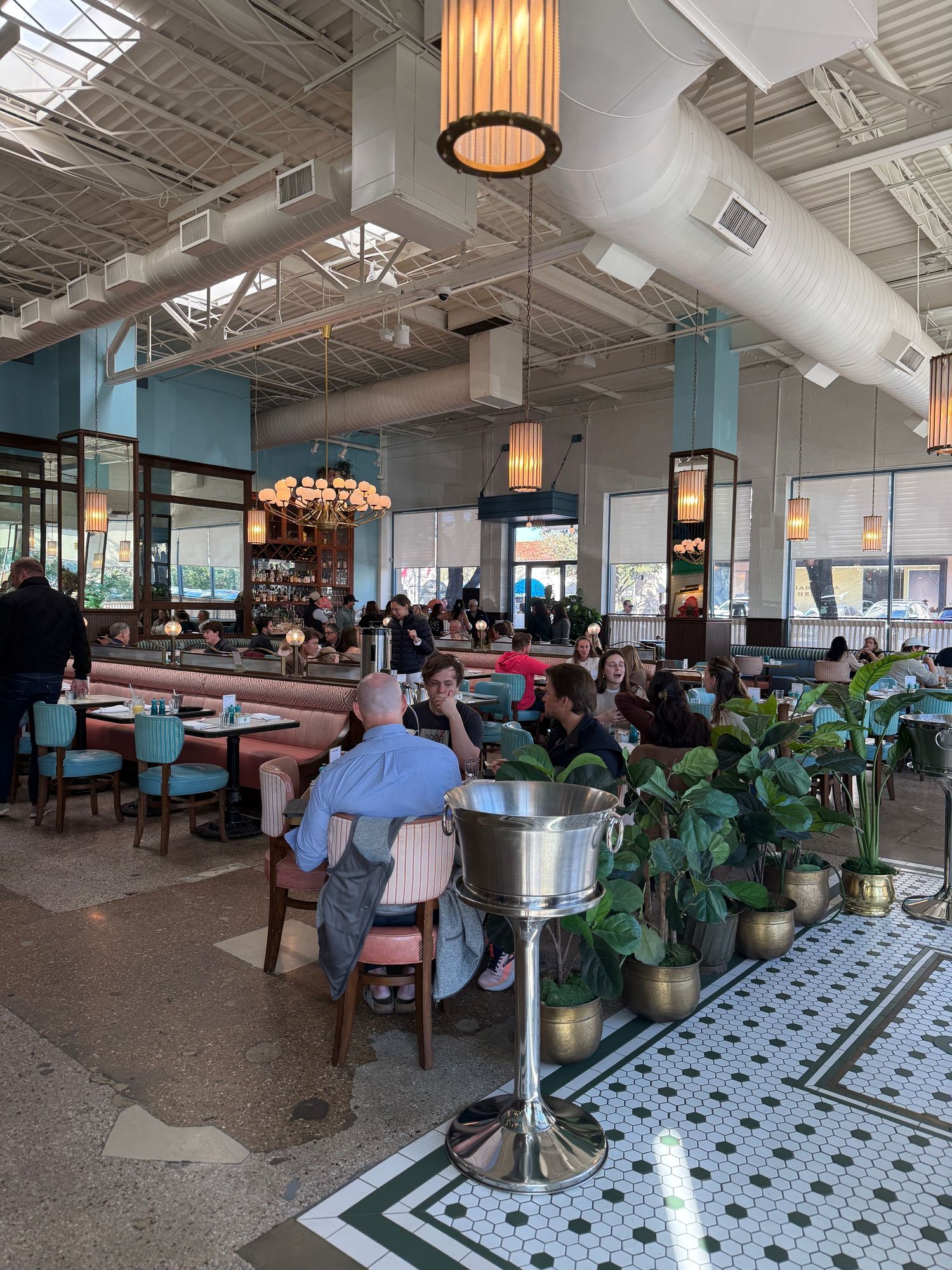 Modern restaurant interior with white tiled floors, hanging pendant lights, and abundant green plants throughout the space.