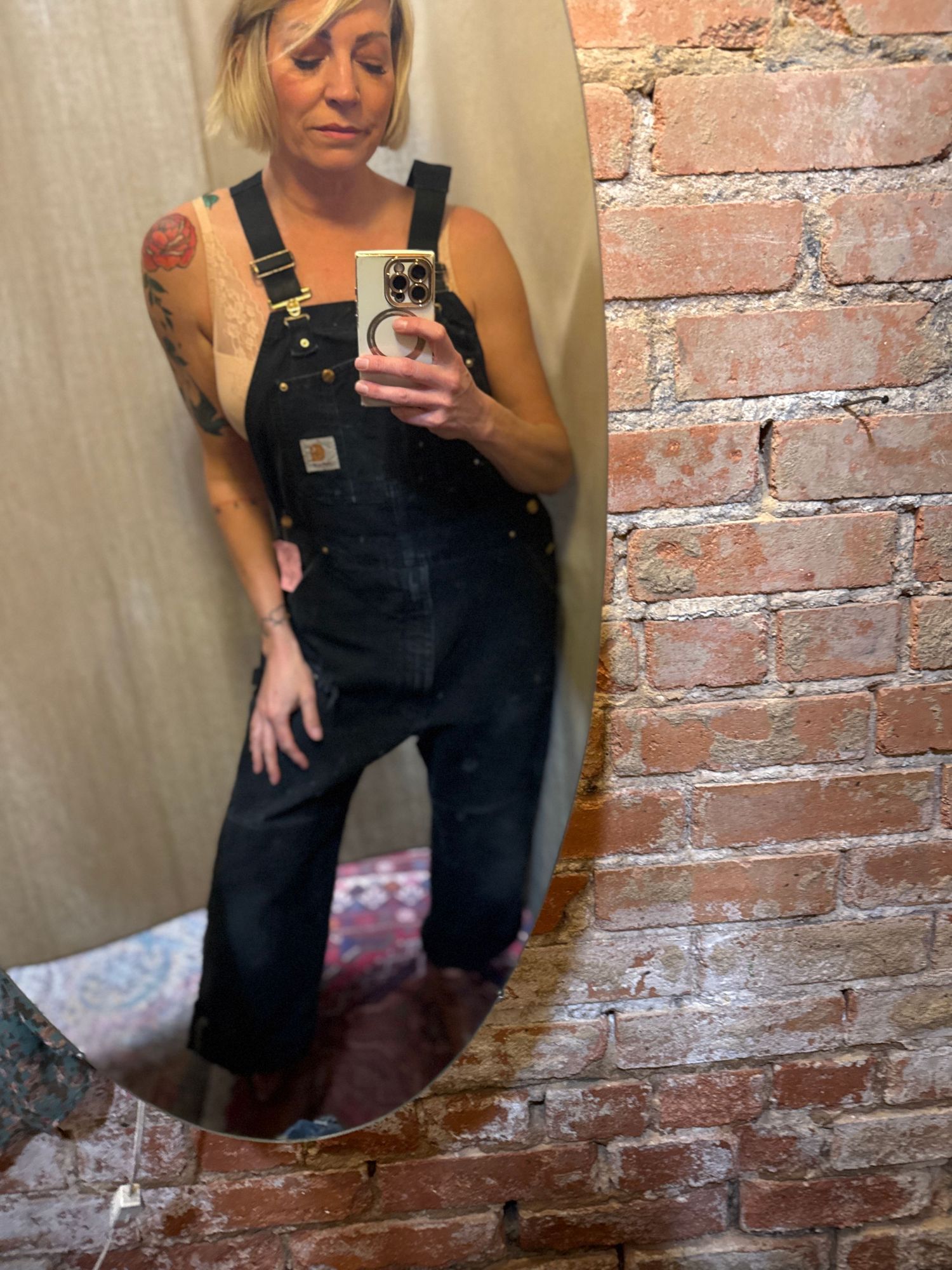 Mirror selfies showing someone wearing black denim overalls against an exposed brick wall backdrop.