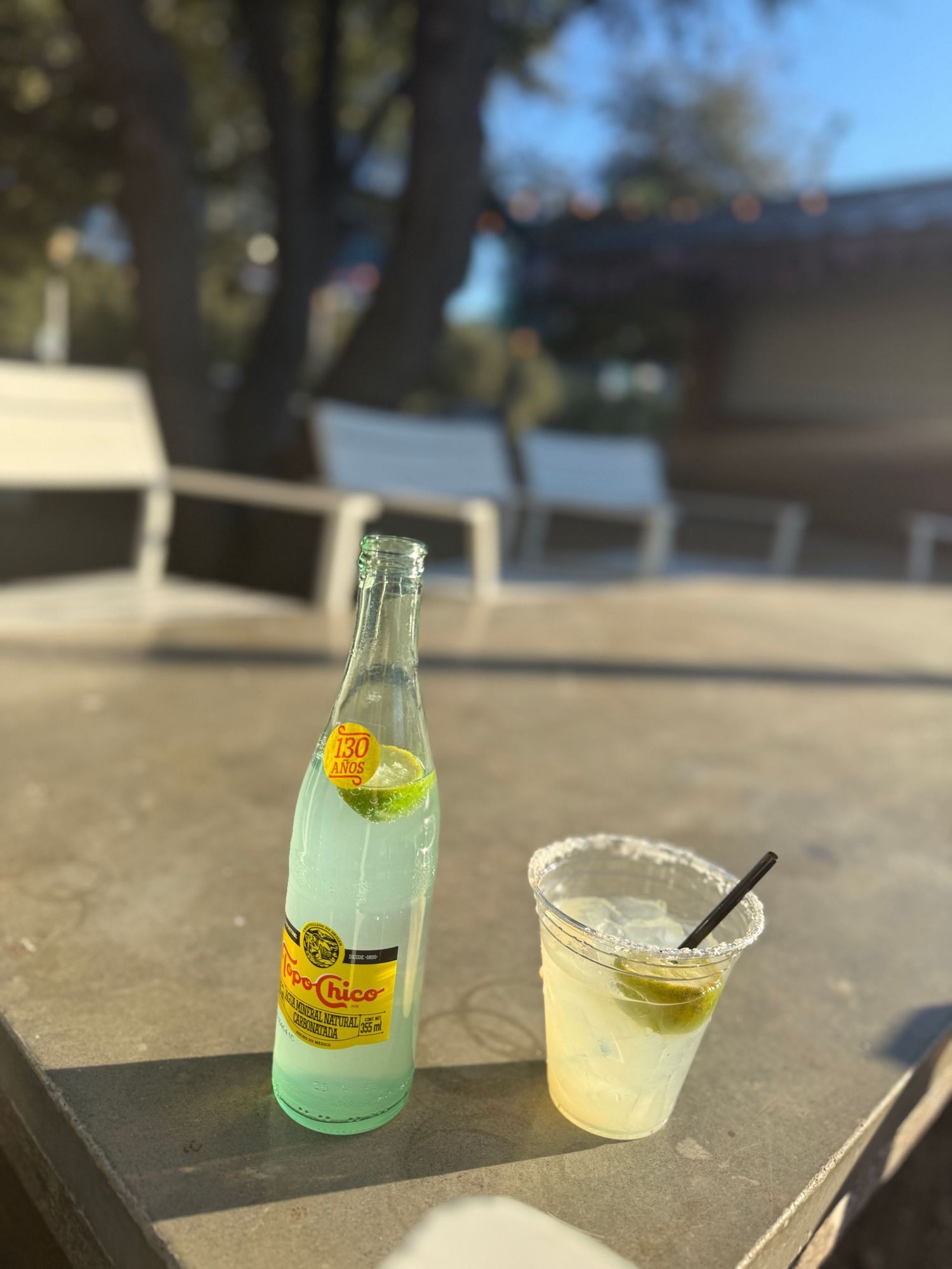 A bottle of Topo Chico and margarita cocktail sit on an outdoor patio table in sunlight.