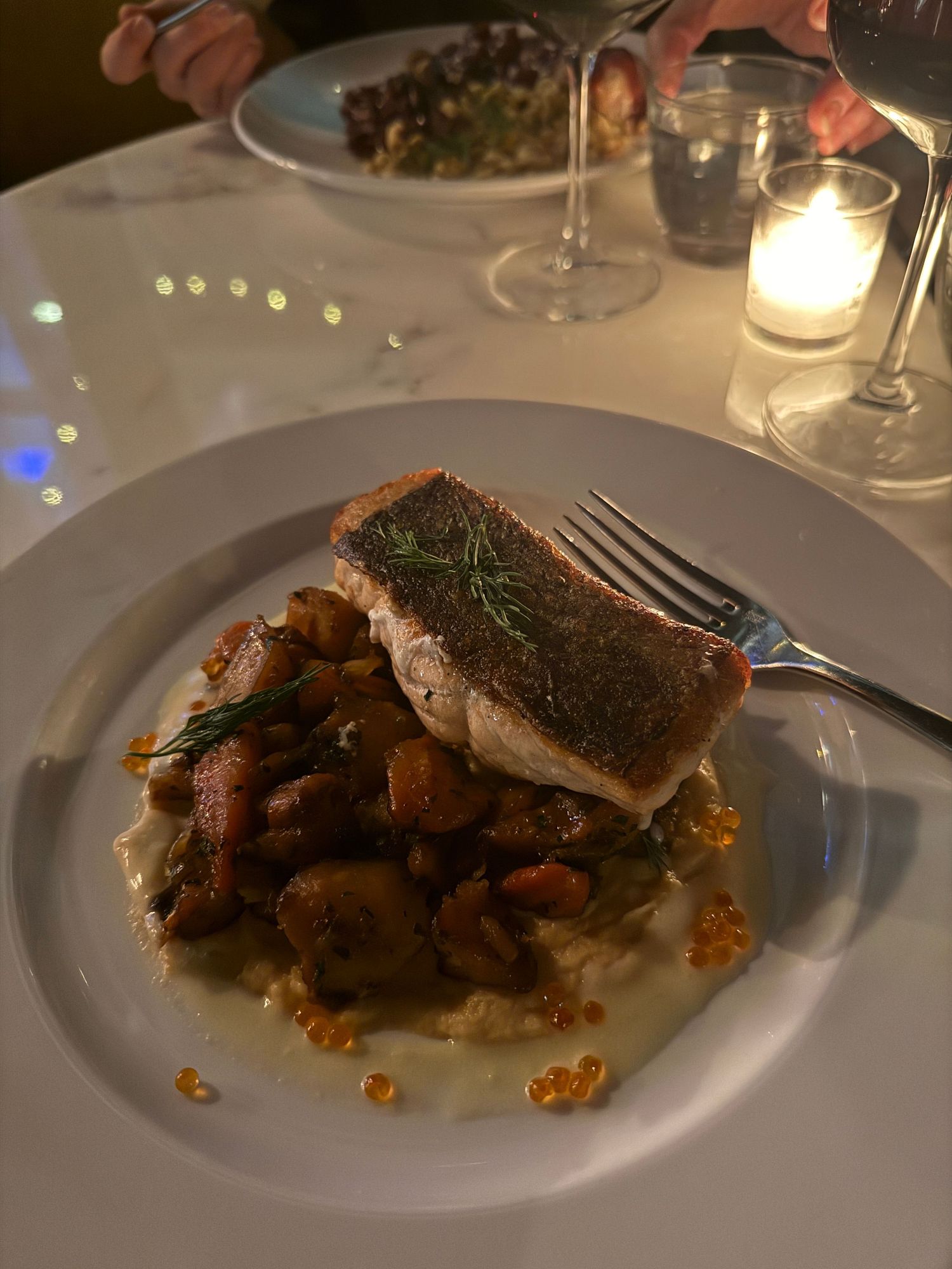 Pan-seared fish fillet served on a bed of roasted vegetables and sauce on an elegant white plate in dim restaurant lighting.
