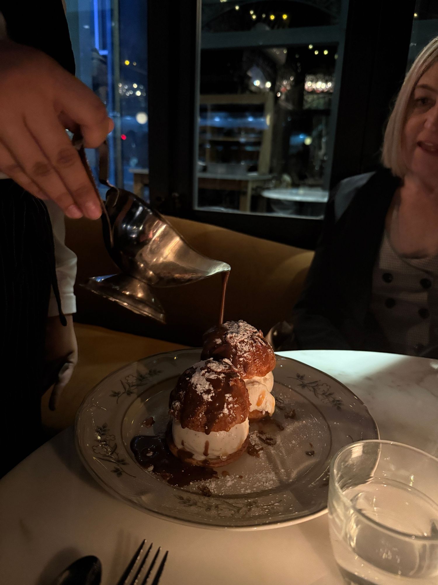 Someone pours chocolate sauce over an elegant dessert of profiteroles dusted with powdered sugar at a dimly lit restaurant.