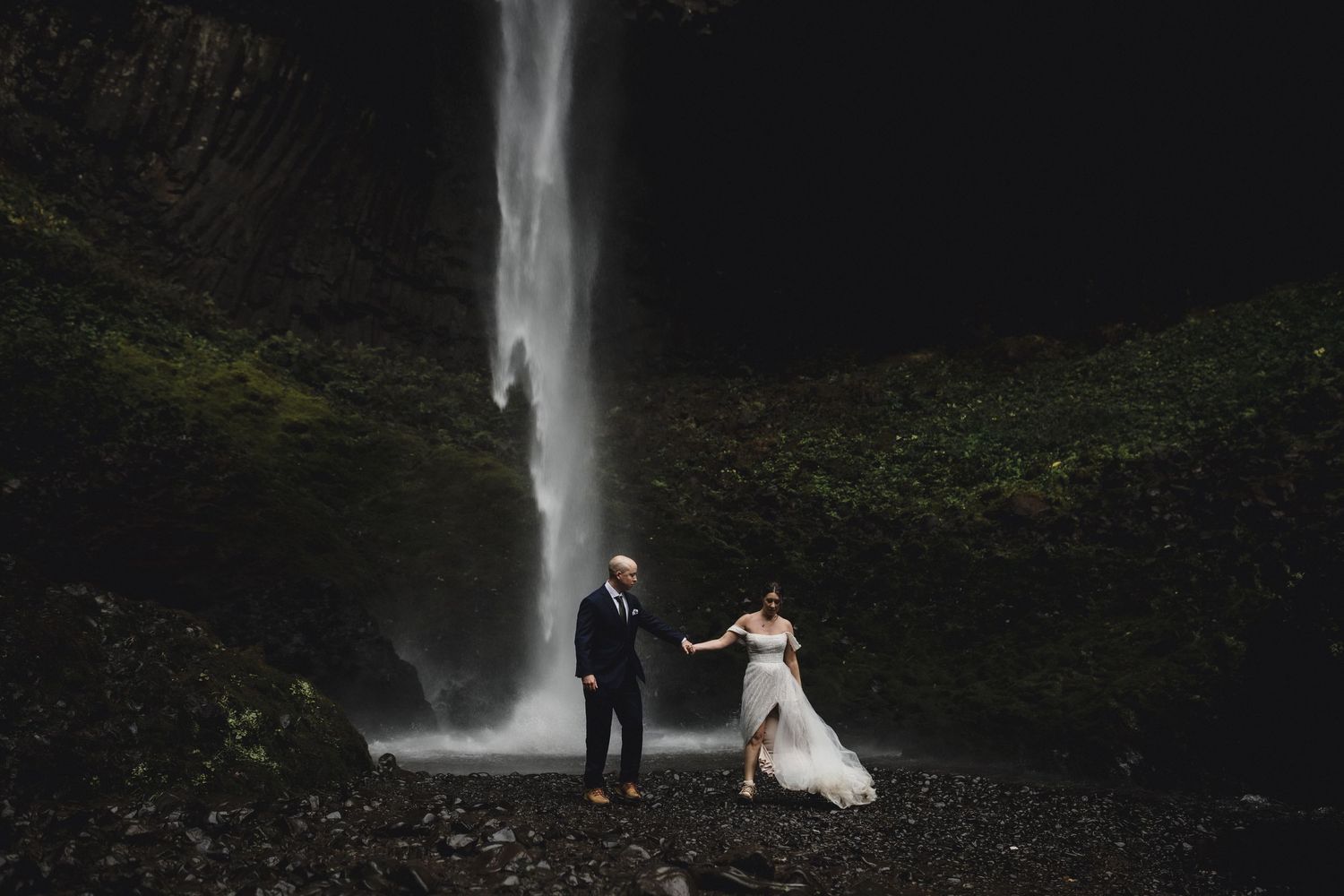 Bride and groom hold hands before a dramatic waterfall in a lush, dark forest during their elopement.