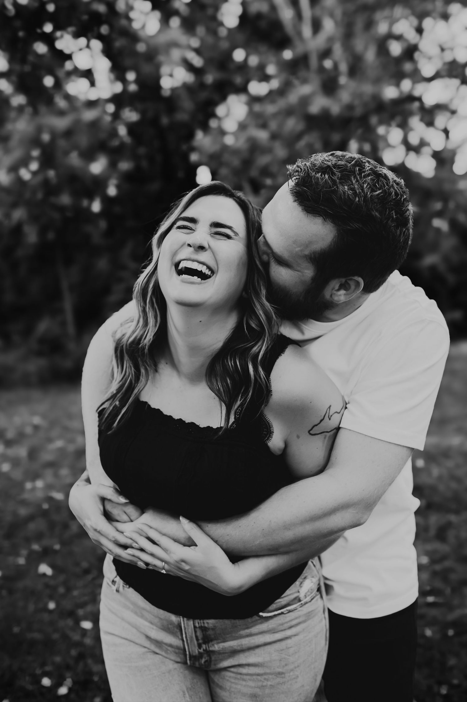 Black and white photo of a laughing woman being hugged and kissed by a man outdoors in a park setting.