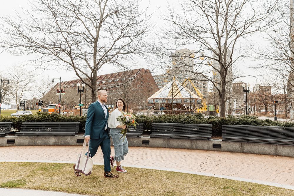 Couple walks hand-in-hand through an urban park in winter, man in blue suit, woman holding white bouquet.